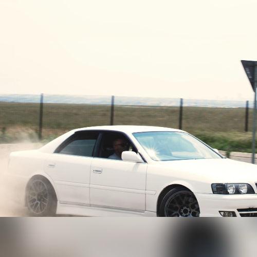 White sports sedan drifting on a dusty track, driver visible through the window with a fence and open field in the background.
