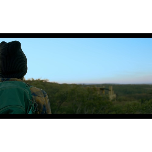 Person with a backpack and beanie gazes at a distant treeline and horizon under a clear blue sky.