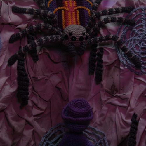 Knitted spider decoration with long black legs and a purple body on wrinkled pink fabric, a smaller purple crochet figure in front.