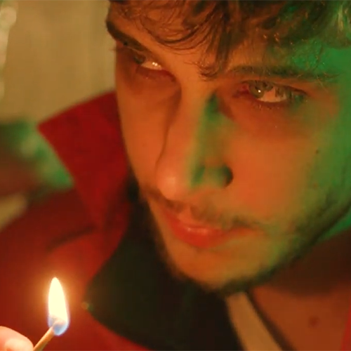 Close-up of a young man with a short beard watching a lit match flame, warm orange light on his face with green lighting nearby.