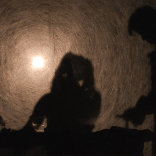 Two people as dark silhouettes appear to kiss, with a bright circular light behind them in a dim setting.