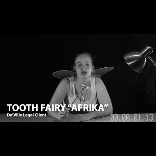 Black-and-white video still of a woman with small fairy wings seated at a desk, holding a wand; on-screen text shows 'TOOTH FAIRY AFRIKA' and a timer reads 00:01:13.