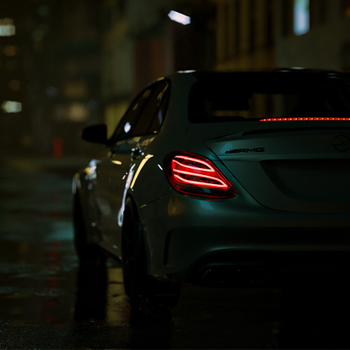 Rear three-quarter view of a white Mercedes-Benz AMG sedan with illuminated taillights on a wet urban street at night.