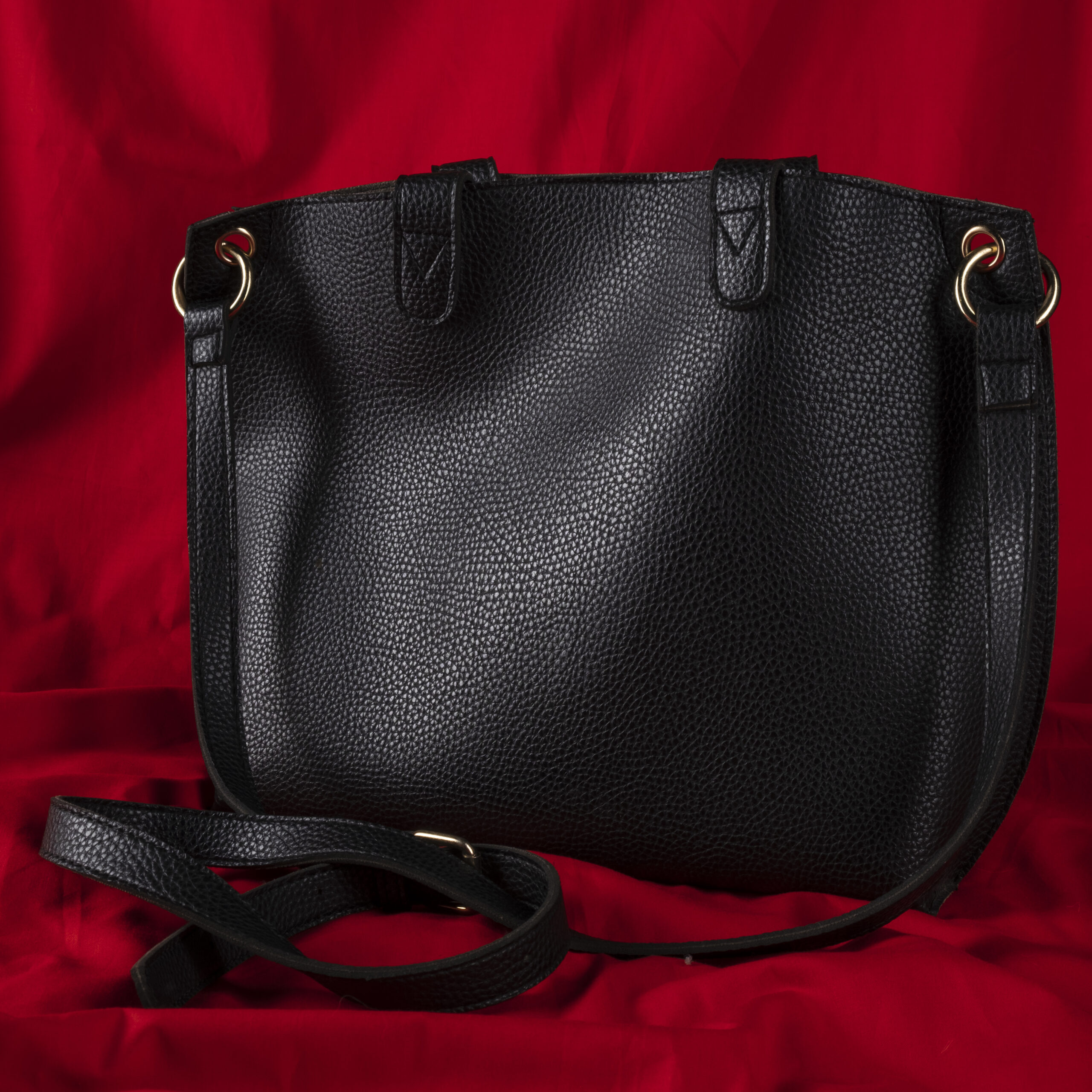 Close-up of a black pebbled leather handbag with gold rings, laid on red fabric, showing straps and texture.