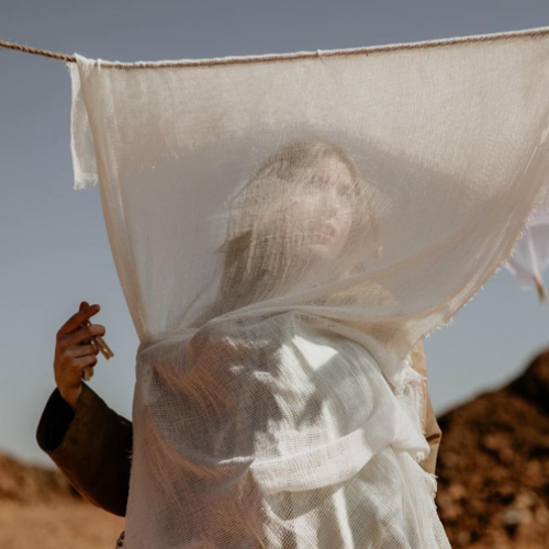 Person wrapped in a sheer white veil over head and shoulders in a desert, with a second hand holding the veil on the left.