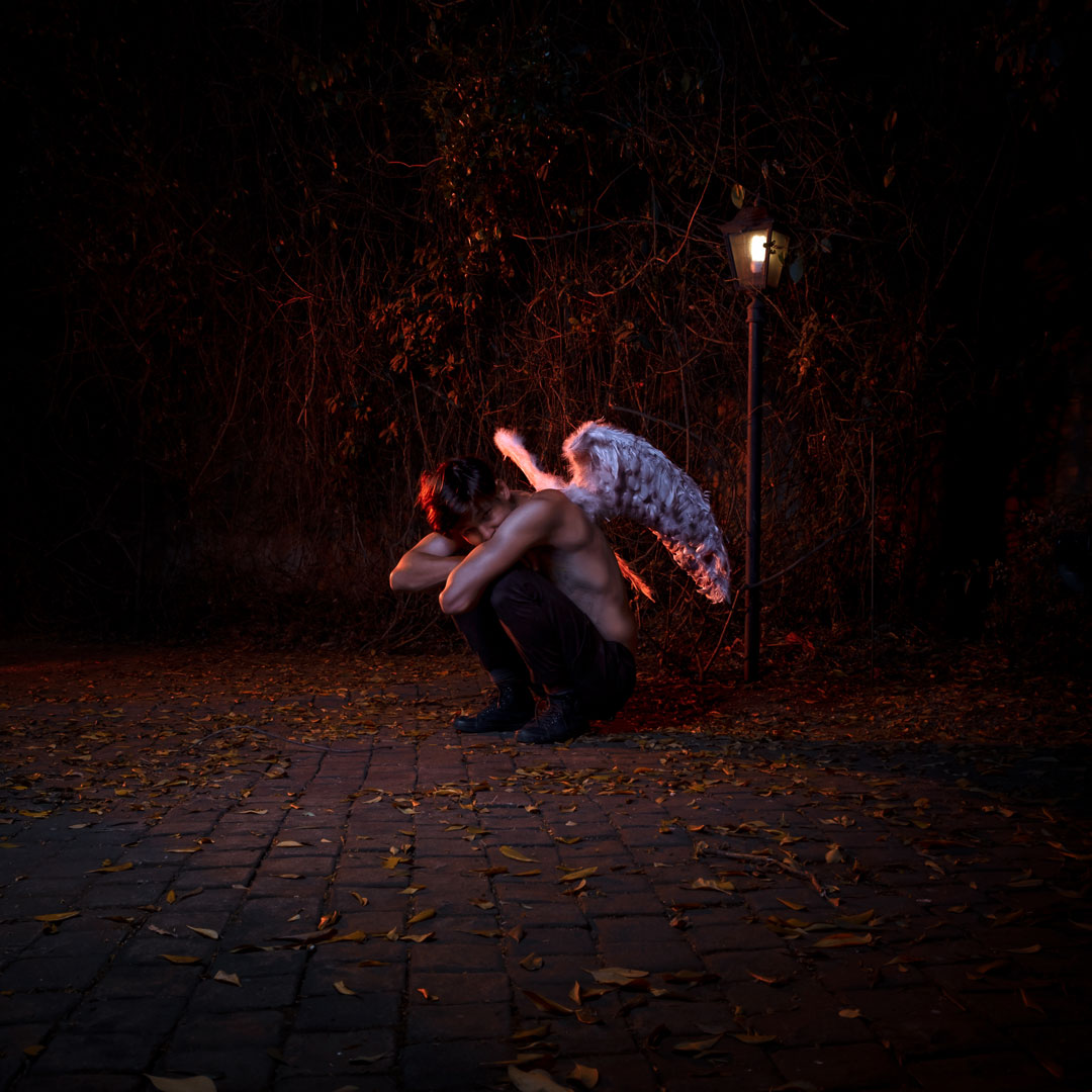 Bare chested person crouches on a cobblestone path at night, white angel wings attached to their back beside a lamppost.