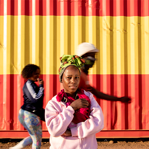 Young girl in a pink jacket and patterned headwrap stands in front of a red-yellow striped shipping container.