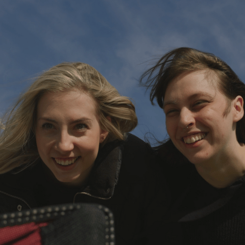 Two friends posing for a selfie outdoors on a sunny day, smiling at the camera with wind-blown hair and a phone visible in the foreground.