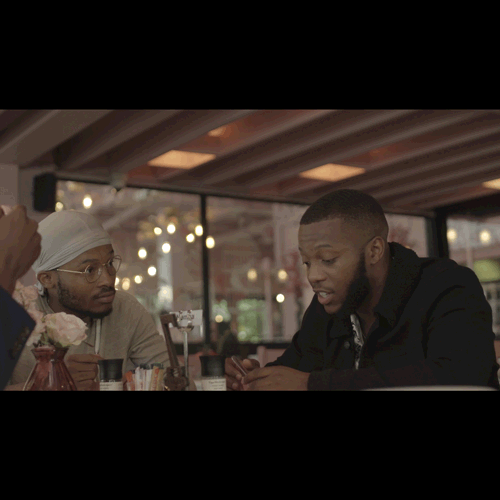 Two men sit at a restaurant table in a cozy cafe, one wearing a white turban and glasses listening to the other speak.