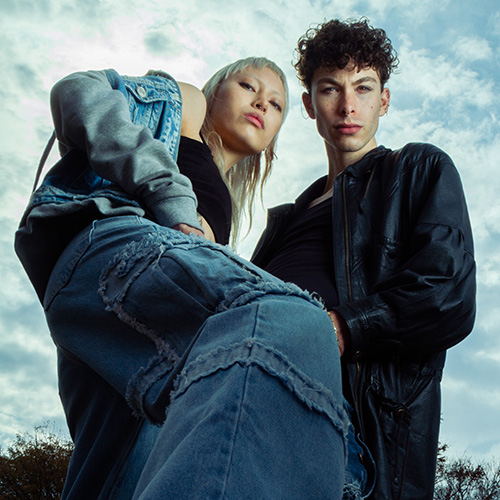 Two young models pose outdoors in denim outfits, shot from a low angle against a cloudy sky.