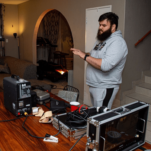 Bearded man in a gray hoodie adjusting a home music setup with a mixer, cases, and cables on a wooden table.