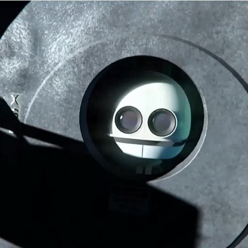 Close-up of a small round robot face with two circular eyes peering through a circular window in a metallic surface.