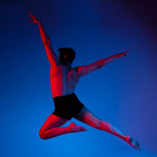 Male dancer mid-leap in blue-red lighting, arms extended, wearing black shorts against a dark studio backdrop
