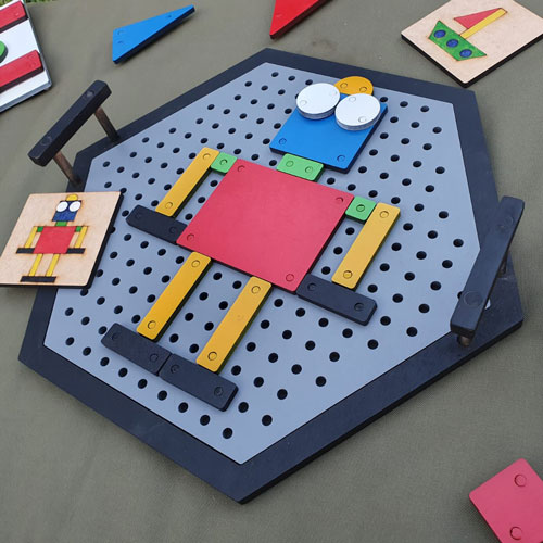 Colorful wooden construction toy assembled on a perforated pegboard to form a small robot with a red square body, yellow limbs, blue head and white eyes.