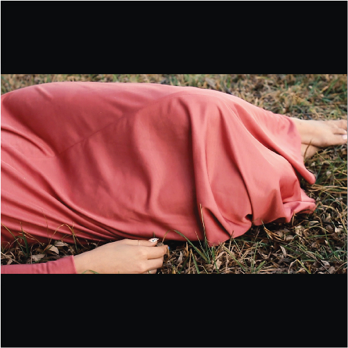 Person lying on the ground outdoors, covered with a pink blanket, with a hand visible at the edge of the cloth.