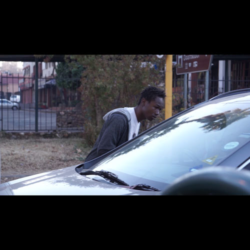 Young man in a gray hoodie leans toward a silver car, looking at the windshield in a parking lot.