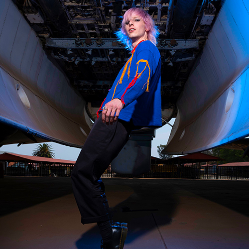 Person with pink and blue hair posing under a large airplane fuselage in a hangar.