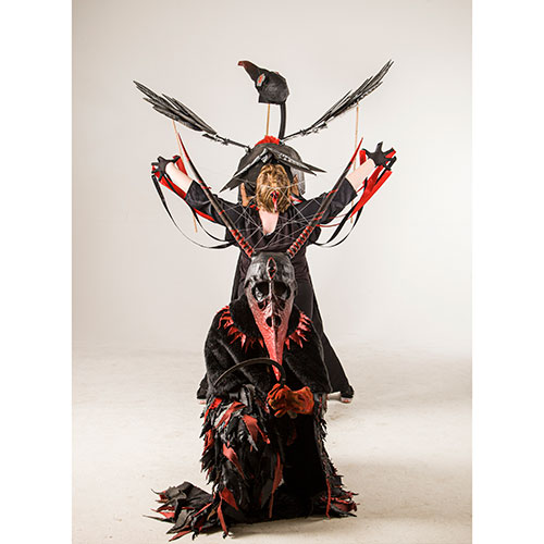Performer in a black-and-red ceremonial costume with a skeletal mask kneels, arms raised with red ribbons, a large black bird prop hovering above.] , but we must ensure proper punctuation and no stray brackets.