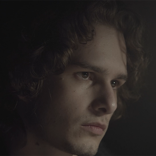 Close-up of a young man with wavy brown hair, looking pensively to the right, lit softly from the left in moody lighting.