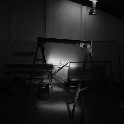 Dimly lit workshop with a large folding sawhorse frame under a bright work light, a bottle resting on the frame, and tools scattered around.