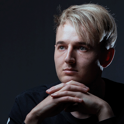 Portrait of a young man with blond hair, resting his chin on clasped hands and looking pensively to the left.