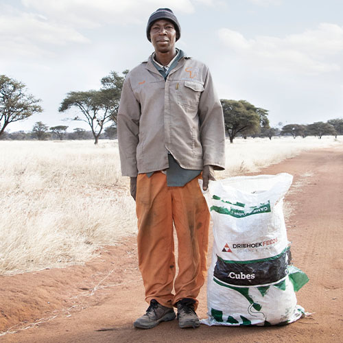 Man in a gray jacket and orange pants stands beside a large white bag labeled 'Cubes' on a red dirt road in a dry savanna.