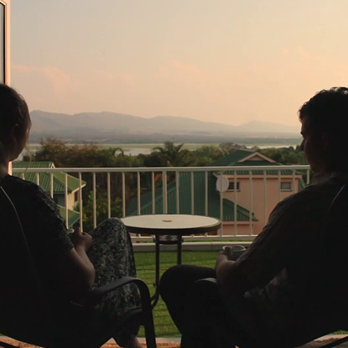 Two people sit on a balcony at sunset, facing a small round table with a railing and a view of houses, greenery, water, and distant hills.