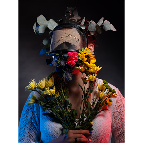 Person wearing a welding helmet with a note reading 'Fuck Society', holding a bouquet of sunflowers and other flowers against a dark studio backdrop.
