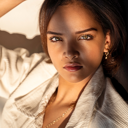 Close-up portrait of a young woman with sunlit skin, wearing a striped jacket and gold jewelry, gazing at the camera with a calm expression.