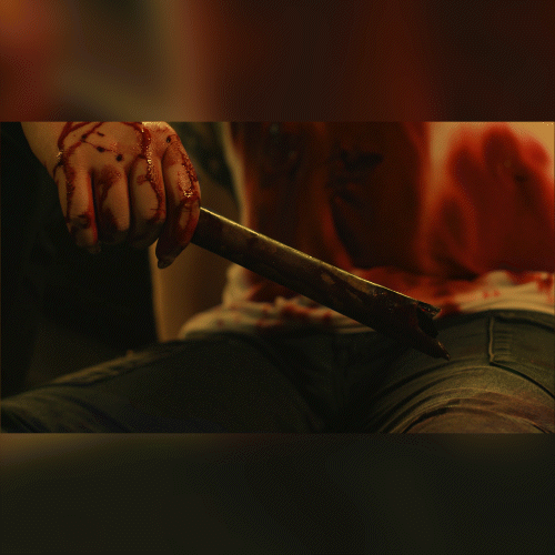 A bloodied hand grips a knife resting on a person’s lap; blood stains cover the hand and clothing.