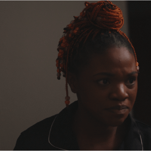Woman with orange braided hair looking serious or contemplative, indoors in low light.