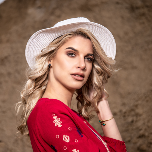 Portrait of a woman with blond wavy hair wearing a white hat and red embroidered top, outdoors against a rocky background.