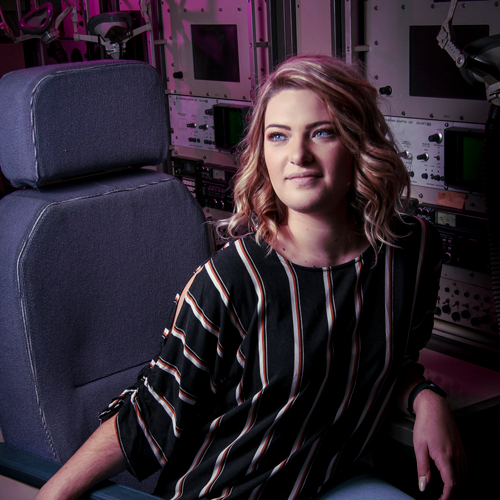 Woman seated in a high-tech control room or cockpit beside equipment panels, looking thoughtful into the distance.
